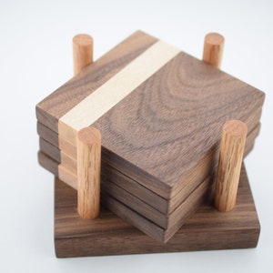 Puede incluir: Un juego de seis posavasos de madera con una incrustación de madera clara, apilados en un soporte hecho del mismo tipo de madera.