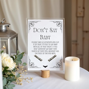 May include: A clear acrylic sign with the text "DON'T SAY BABY" and instructions for a baby shower game. The sign includes decorative flourishes, clothespin illustrations, and a wooden base. A lit candle and floral arrangement are also in the image.