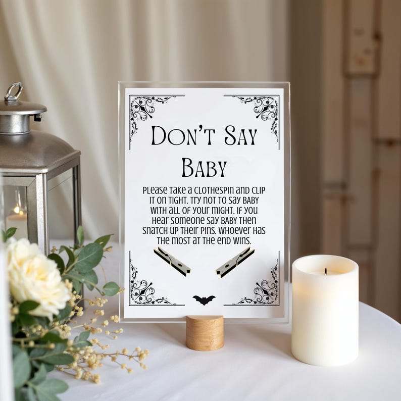 May include: A clear acrylic sign with the text "DON'T SAY BABY" and instructions for a baby shower game. The sign includes decorative flourishes, clothespin illustrations, and a wooden base. A lit candle and floral arrangement are also in the image.