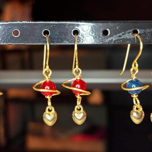 May include: Five gold-toned earrings featuring a central spherical bead encircled by a thin gold wire ring, resembling a planet with rings. Each earring includes a small gold-toned heart charm dangling at the bottom and a curved ear wire hook.