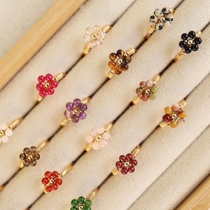 May include: A collection of gold-toned wire rings, each adorned with a cluster of colorful beads arranged in a flower shape. The beads vary in color, including red, purple, pink, and brown. The rings are displayed in a beige jewelry box.