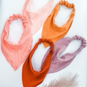 Op de afbeelding: Vier stoffen haarbanden in verschillende kleuren: perzik, oranje, roest en mauve. De haarbanden zijn gemaakt van een zachte, textuurrijke stof en hebben een geplooide elastische band.