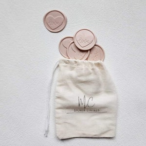 May include: A small white drawstring bag with the text "MC SILVER LININGS" printed on the front. The bag is filled with pink heart-shaped wax seals with the word "love" inscribed on each.
