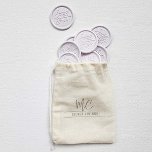 May include: A small white drawstring bag with the text "MC SILVER LININGS" printed on the front. The bag is filled with lavender-colored wax seals with the text "Just the Two of Us" embossed on them.