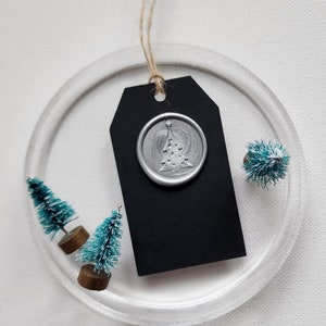 May include: A clear, circular ornament with a black tag and a silver wax seal featuring a Christmas tree design. Miniature teal bottle brush trees are arranged around the tag. A natural twine loop is attached for hanging. Festive holiday decor.