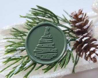 May include: A green wax seal with a stylized Christmas tree design. The seal is on a white background with a pine branch and a pine cone.
