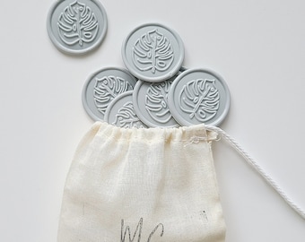 May include: A small white drawstring bag with the text "MC SILVER LININGS" printed on the front. The bag is filled with gray wax seals featuring a leaf design.