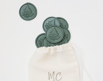 May include: A white drawstring bag with the text "MC SILVER LININGS" printed on the front. The bag is filled with green wax seals with a fern design.