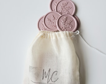 May include: A small white cloth bag with a drawstring closure. The bag has a black logo that says "MC SILVER LININGS". The bag is filled with pink wax seals with a leaf design.
