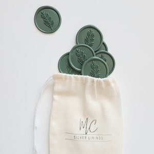 May include: A small white drawstring bag with the text "MC SILVER LININGS" printed on it. The bag is filled with green wax seals with a leaf design.
