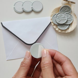 May include: A white envelope with a black border and a gray wax seal with a white adhesive backing. The seal is being held by a hand. There are several other gray wax seals in a seashell with the text "Thank you" on them.