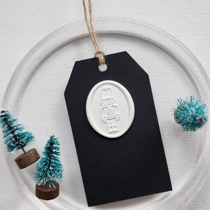 May include: A black gift tag with a white wax seal featuring a nutcracker design, suspended by a natural twine string. Miniature teal Christmas trees and a clear acrylic ring surround the tag, creating a festive holiday theme.