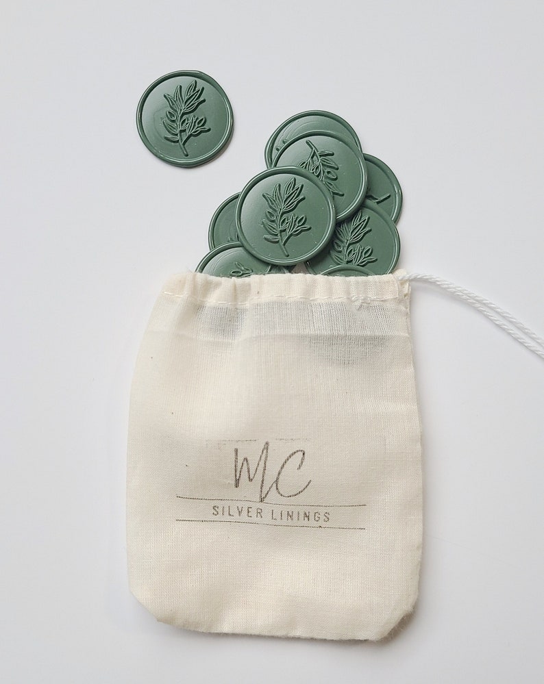 May include: A small white drawstring bag with the text "mc SILVER LININGS" printed on the front. The bag is filled with green wax seals with a leaf design.