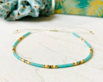 May include: A delicate bracelet made with tiny turquoise and white beads, accented with gold beads. The bracelet is strung on a white cord and has a simple knot closure.