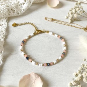 May include: A delicate bracelet with a gold chain and clasp. The bracelet features a pattern of white, pink, and dark blue beads, interspersed with small gold-colored beads. The bracelet is displayed on a white surface with lace and flowers.