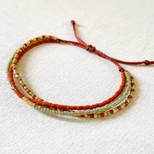 Puede incluir: Una pulsera hecha a mano con tres hilos de pequeñas cuentas de colores. Los hilos son en tonos de rojo, verde y dorado, ensartados en un cordón marrón. La pulsera es ajustable y se ata con un nudo.