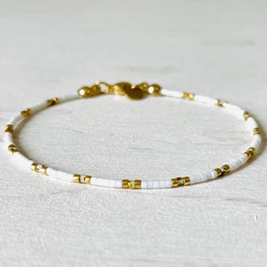Peut inclure: Bracelet de perles blanches délicat avec des accents dorés. Le bracelet présente de petites perles blanches carrées entrecoupées de perles dorées. Un fermoir doré sécurise le bracelet.