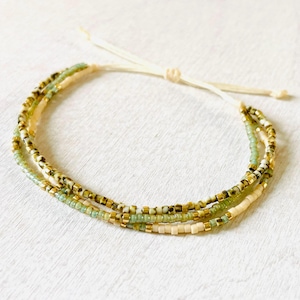 Könnte beinhalten: Mehrsträngiges Armband mit grünen, goldenen und cremefarbenen Perlen. Das Armband hat eine verstellbare Kordel und einen Knotenverschluss in Creme. Die Perlen sind klein und gleichmäßig verteilt.