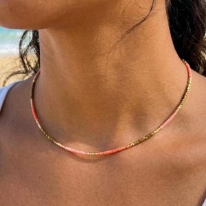 Peut inclure: Un collier de perles délicat avec un dégradé de couleurs. Le collier présente de petites perles rondes dans des tons or, corail et rose. Le collier est porté autour du cou et est un accessoire simple et élégant.