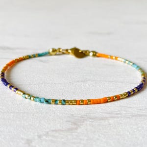 Peut inclure: Un bracelet délicat en or avec un motif de perles multicolores. Les perles sont disposées en dégradé de violet, turquoise et orange.