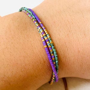 Puede incluir: Un brazalete de múltiples hilos con cuentas moradas, turquesa y doradas. El brazalete está hecho de pequeñas cuentas cuadradas y se lleva en la muñeca.