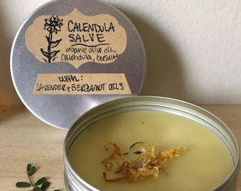 May include: A silver tin with a lid containing a yellow salve with dried flower petals. The lid has a brown paper label with the text "Calendula Salve" and "organic olive oil, calendula, beeswax" and "with: lavender + bergamot oils".