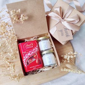 May include: A gift box containing a red Lindor chocolate box, two small glass jars of candles, and a separate gift box tied with a ribbon. The candles are labelled "Cinnamon Spice" and "Pomegranate Spice". A "Thank you" tag is attached.