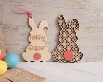 May include: Two wooden Easter bunny decorations. One bunny has the words "happy Easter" cut out, a pink tail, and a pink ribbon for hanging. The other bunny has a geometric pattern cut out and a pink tail. Easter decor.