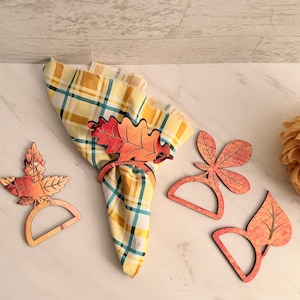 May include: A collection of fall-themed napkin rings and a plaid napkin. The napkin rings are shaped like leaves in shades of orange, red, and brown. The napkin has a yellow, blue, and white plaid pattern. A large, yellow flower is in the background.