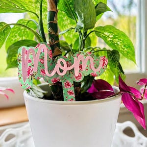 May include: A white potted plant with green and pink foliage. A decorative plant stake with the word "Mom" in pink script is inserted into the soil. The stake has a floral pattern with a mint green background.