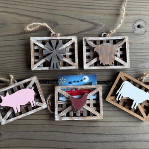 May include: A collection of rustic wooden ornaments with farm animal and windmill designs. Each ornament features a different animal or object, including a pig, cow, chicken, and windmill, set within a wooden frame. They are strung with twine for hanging.