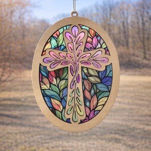 May include: Wooden oval-shaped stained glass cross with a suction cup for hanging. The cross features a colorful leaf design in shades of pink, purple, green, and blue. The wooden frame has a natural wood finish.