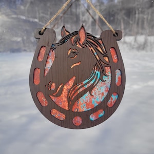 May include: Wooden horse head and horseshoe wall hanging. The dark brown wood features a cutout design of a horse's head within a horseshoe shape. The interior has a distressed orange and teal color scheme, with a natural twine hanger.