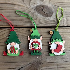 May include: Three wooden Christmas gnome ornaments with green hats, white beards, and red and green accents. Each ornament has a different design: a Christmas tree, a gift, and a stocking. They have red, green, and yellow hanging ribbons.