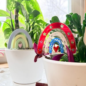 Puede incluir: Dos plantas en macetas blancas con decoraciones en forma de arcoíris. Un arcoíris es rojo, blanco y azul con recortes de estrellas, y el otro es pastel con un corazón rosa. Plantas verdes y una ventana en el fondo.