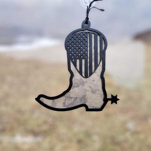 May include: Black and gray cowboy boot-shaped ornament with an American flag design. The top portion features the stars and stripes, while the boot has a textured, marbled appearance. A spur is visible.