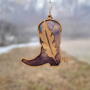 May include: Wooden ornament shaped like a cowboy boot with a feather design. The boot is brown with a purple and blue background. A gold-colored feather is layered over the boot. The ornament is suspended by a natural-colored string.