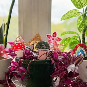 May include: A collection of potted plants with decorative accents. The plants include a snake plant, a plant with large green leaves, and a plant with purple foliage. The decorations include mushroom, flower, bird, and rainbow shapes.
