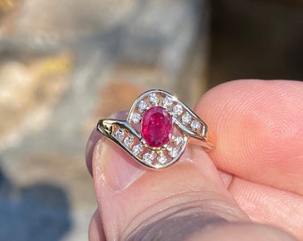 以下が含まれることがあります： 赤いルビーの宝石を、小さなダイヤモンドのハローで囲んだ金のリング。リングはユニークな曲線デザインです。