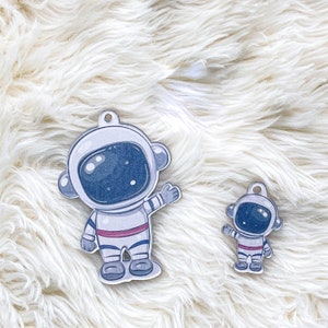May include: Two white astronaut ornaments with blue details. The larger astronaut has a raised arm and is wearing a red and white striped belt. The smaller astronaut has both arms down.