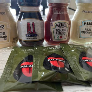 May include: An assortment of condiment bottles and olive oil packets. The bottles include Heinz Dijon Mustard, A1 Sauce, Heinz Tomato Ketchup, and Heinz Real Mayonnaise. The packets are labeled Paladin Azeite Virgem Extra.