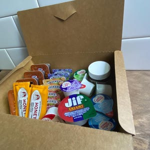 May include: A brown cardboard box filled with individual servings of condiments. The box contains packets of honey, peanut butter, and coffee creamer, along with small containers of jelly and other spreads.