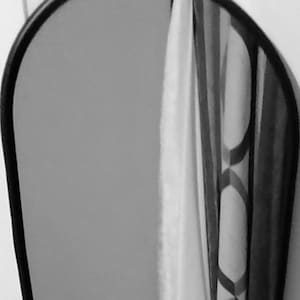 May include: A black oval mirror with a simple ring design at the top. The mirror is hanging on a white wall and reflects a white towel.