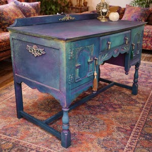 May include: An antique desk, hand-painted in blue, purple, and teal with gold detailing. The desk has decorative carvings, brass hardware, and a tassel. It features a scalloped edge and stands on four turned legs. The desk is a statement piece.
