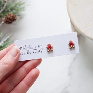 Puede incluir: Pendientes de botón con forma de hombre de jengibre con lazo rojo. Se muestran en una tarjeta blanca con el texto "Ruthie's Art & Clay". Fondo con rama de pino y superficie clara.