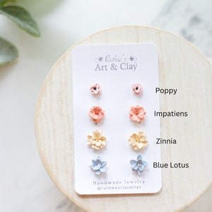 May include: A set of handmade flower stud earrings on a white card. The earrings are in the shape of poppies, impatiens, zinnias, and blue lotus flowers. The card has the text "Ruthie's Art & Clay" and "Handmade Jewelry @ruthiesartandclay".