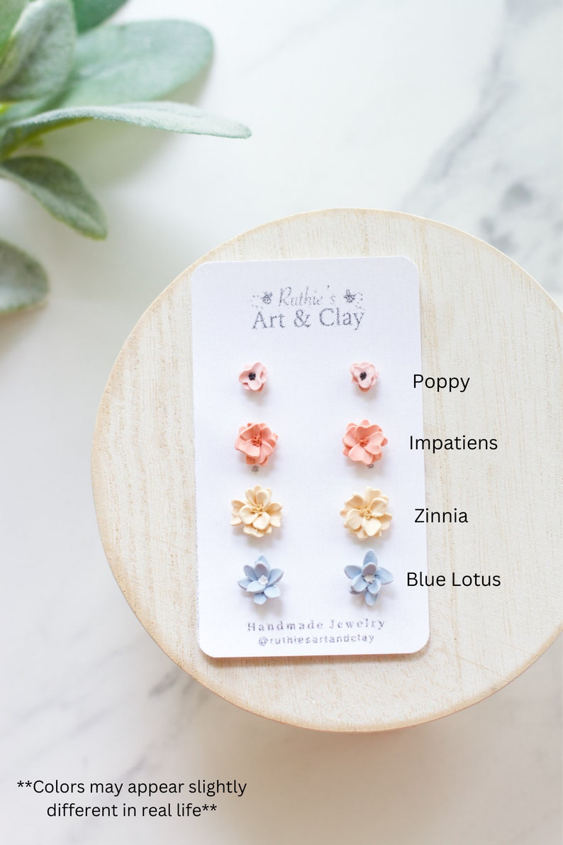 May include: A set of handmade flower stud earrings on a white card. The earrings are in the shape of poppies, impatiens, zinnias, and blue lotus flowers. The card has the text "Ruthie's Art & Clay" and "Handmade Jewelry @ruthiesartandclay".