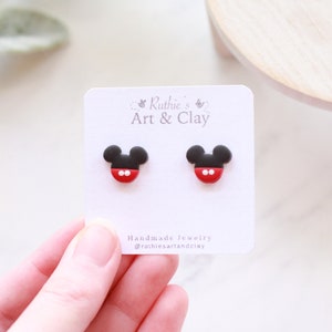 Puede incluir: Un par de pendientes de bot&oacute;n con forma de Mickey Mouse en negro, rojo y blanco. Los pendientes se muestran en una tarjeta blanca con el texto "Ruthie's Art & Clay".