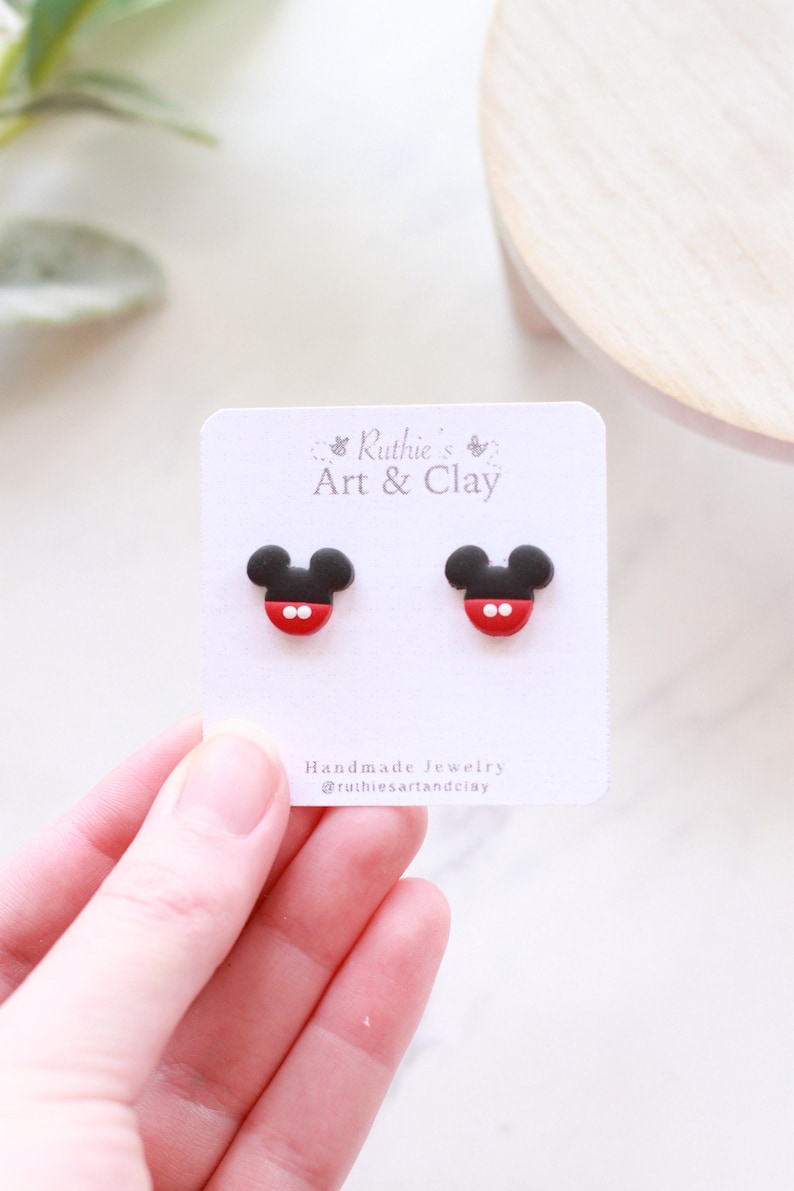 Puede incluir: Un par de pendientes de bot&oacute;n con forma de Mickey Mouse en negro, rojo y blanco. Los pendientes se muestran en una tarjeta blanca con el texto "Ruthie's Art & Clay".