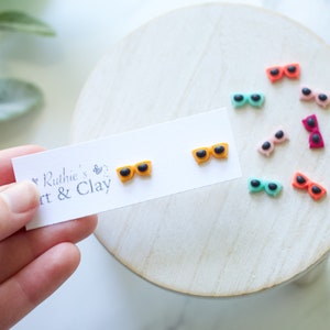 Puede incluir: Pendientes con forma de gafas de sol de varios colores. Dos pares naranjas están sobre una tarjeta blanca con el texto "Ruthie's Art & Clay". Otros pendientes rosas, rojos y turquesas están sobre una superficie de madera.